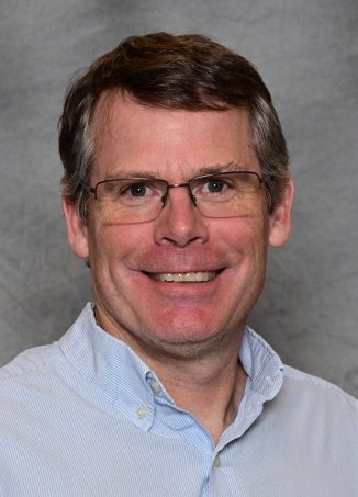 Professional headshot of middle-aged man wearing glasses and light blue shirt.